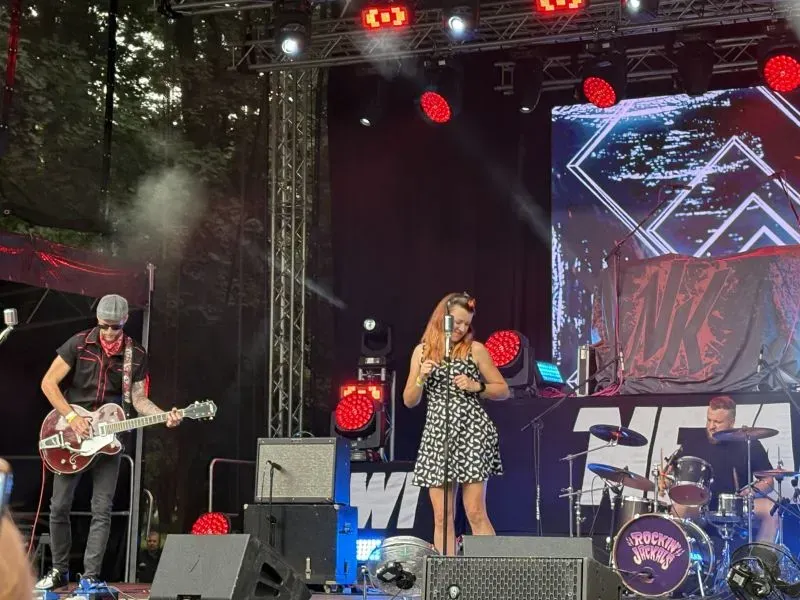 Zespół muzyczny na scenie z gitarzystą, wokalistką i perkusistą podczas koncertu.