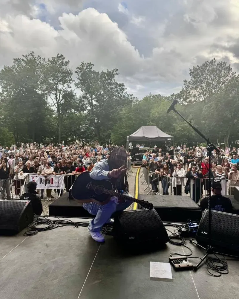 Muzyk z gitarą akustyczną kuca na scenie przed tłumem zgromadzonym na koncercie na świeżym powietrzu.