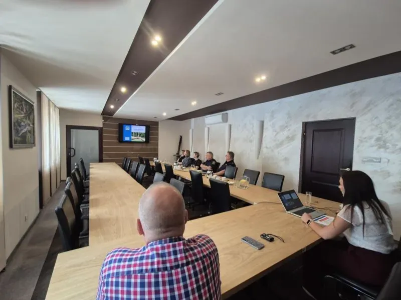 Kilka osób siedzi przy długim drewnianym stole konferencyjnym w sali, na której ścianie wisi telewizor z wyświetlanym obrazem.