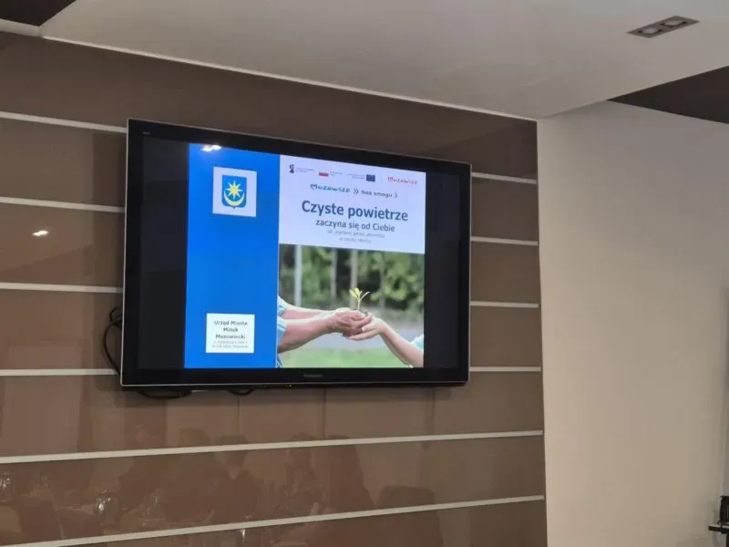 Ekran telewizora ściennego wyświetlający plakat kampanii „Czyste powietrze zaczyna się od Ciebie” z grafiką dwóch rąk trzymających młodą roślinę.