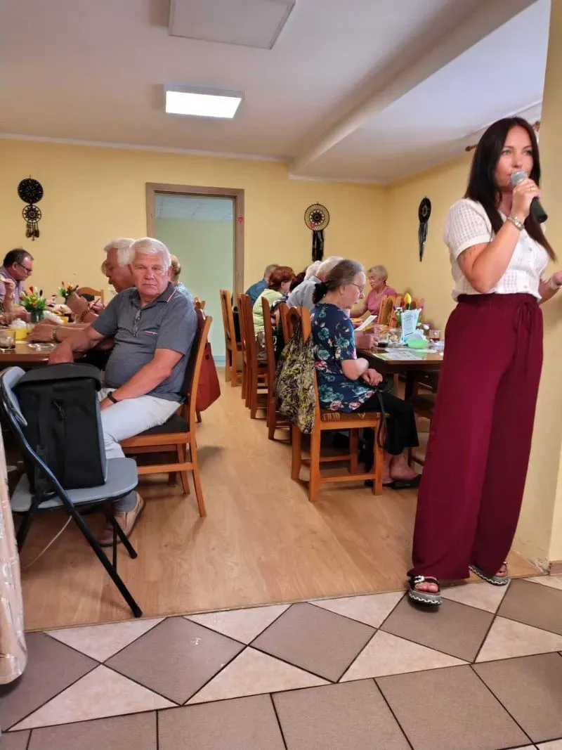 Kobieta mówi do mikrofonu obok stołu, przy którym siedzą starsi ludzie.