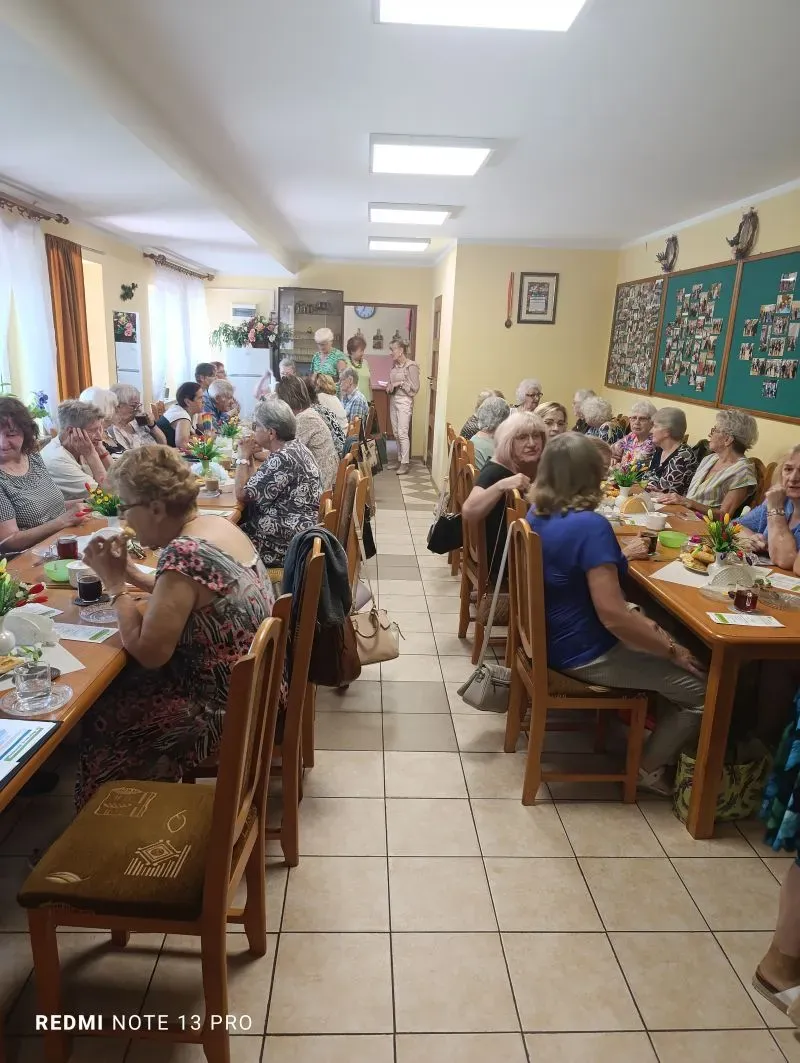 Grupa starszych osób siedzi przy dwóch długich stołach w świetlicy, rozmawiając i jedząc posiłki.
