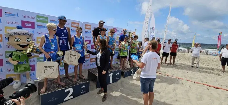 Zawodnicy na podium plażowym odbierający nagrody podczas ceremonii wręczenia pucharów na piasku, obok stoi maskotka i osoby wręczające trofea.