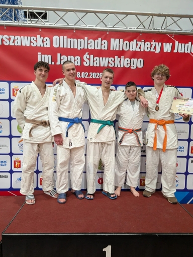 Pięciu młodych zawodników stoi razem na podium w białych judogach z różnokolorowymi pasami. Mają ręce na ramionach, jeden trzyma medal i dyplom; w tle duży czerwony baner i jasne wnętrze hali.