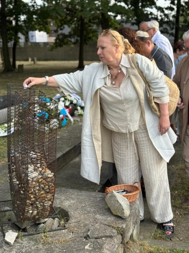 Kobieta w jasnym ubraniu układa kamień na metalowej konstrukcji na zewnątrz, w tle grupa osób oraz zielone drzewa.