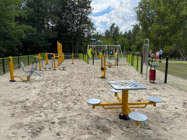Siłownia plenerowa z urządzeniami do ćwiczeń, stołem do gry w szachy i placem zabaw z zjeżdżalnią, otoczona ogrodzeniem i drzewami.