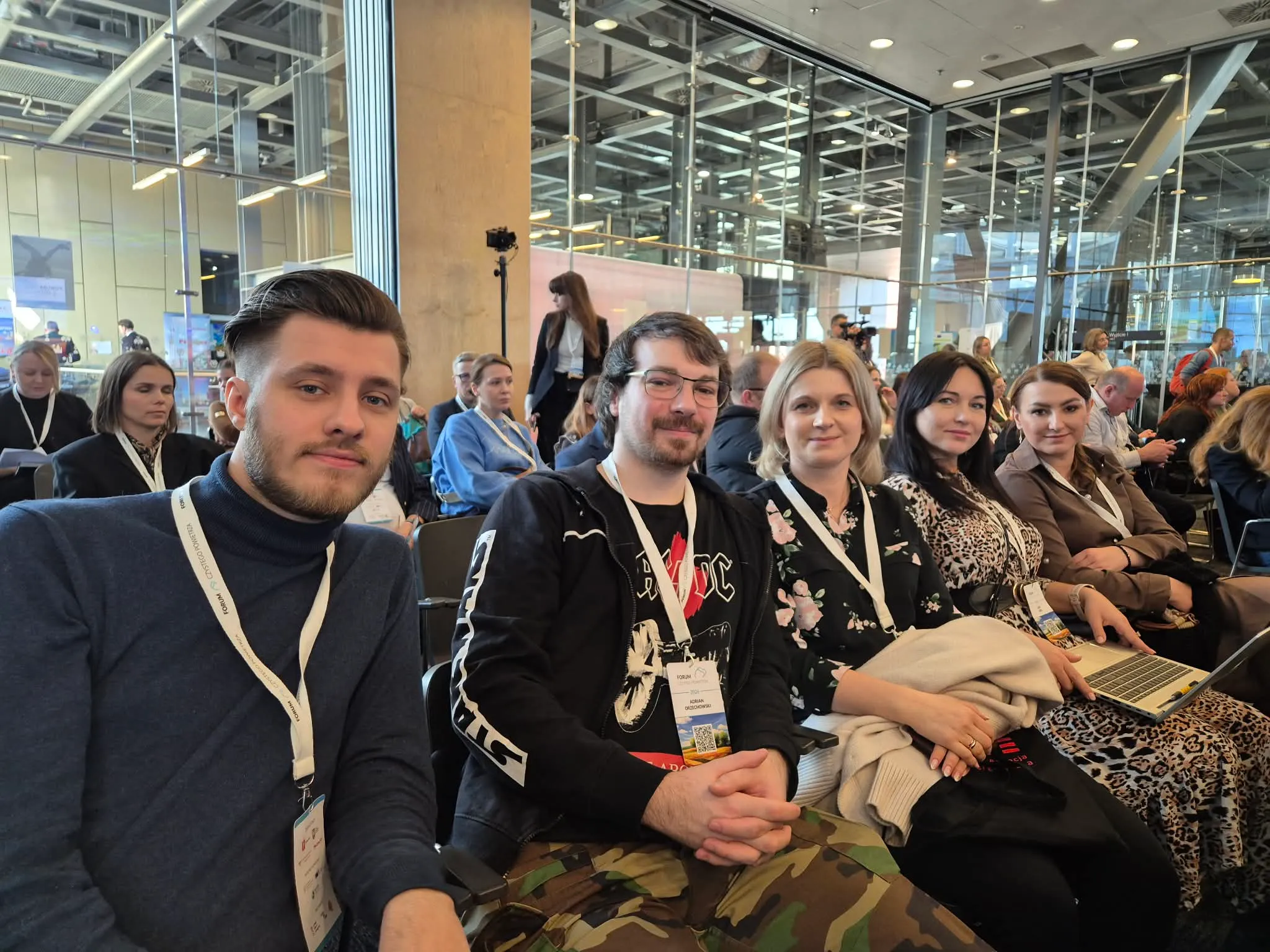 Pięć osób siedzi obok siebie w rzędzie sali konferencyjnej. Wszyscy mają identyfikatory na smyczach. Mężczyźni siedzą z lewej, kobiety po prawej. Kobieta najbardziej po prawej trzyma laptopa na kolanach. Za nimi widoczna jest przeszklona ściana i sufit z metalowymi instalacjami.