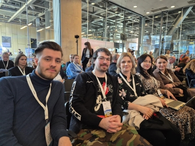 Pięć osób siedzi obok siebie w rzędzie sali konferencyjnej. Wszyscy mają identyfikatory na smyczach. Mężczyźni siedzą z lewej, kobiety po prawej. Kobieta najbardziej po prawej trzyma laptopa na kolanach. Za nimi widoczna jest przeszklona ściana i sufit z metalowymi instalacjami.