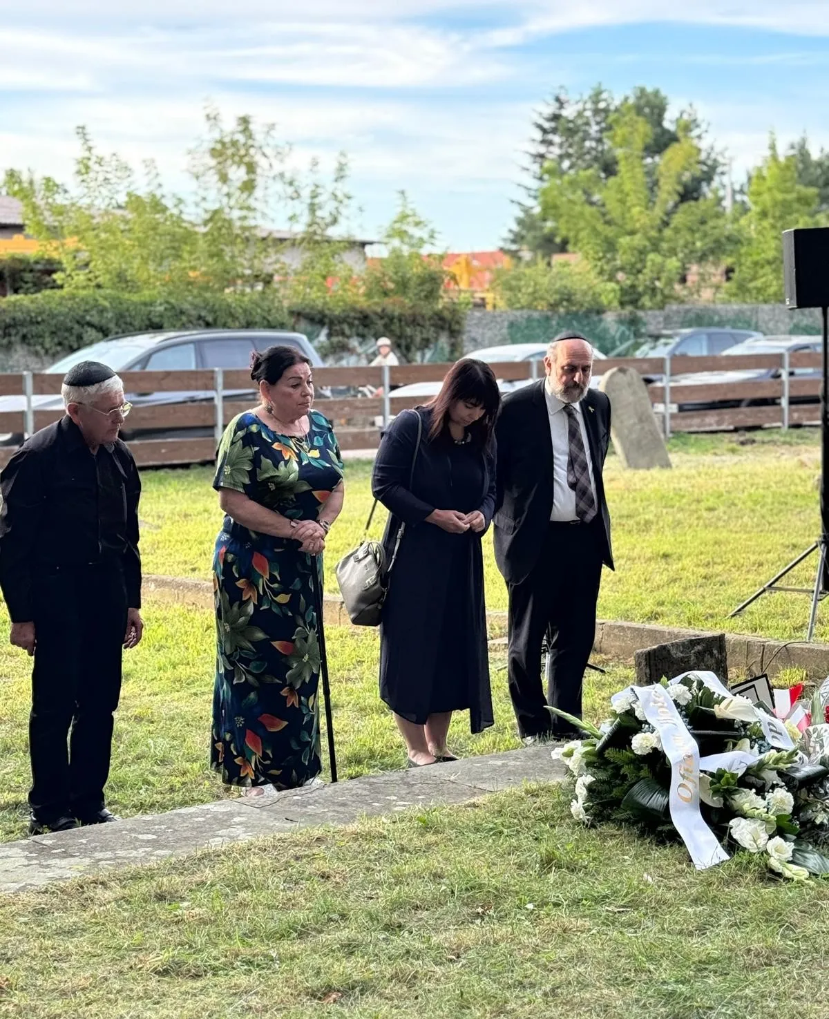 Cztery osoby stoją na zewnątrz nad kwiatami położonymi na ziemi, za nimi widać ogrodzenie, samochody i drzewa.