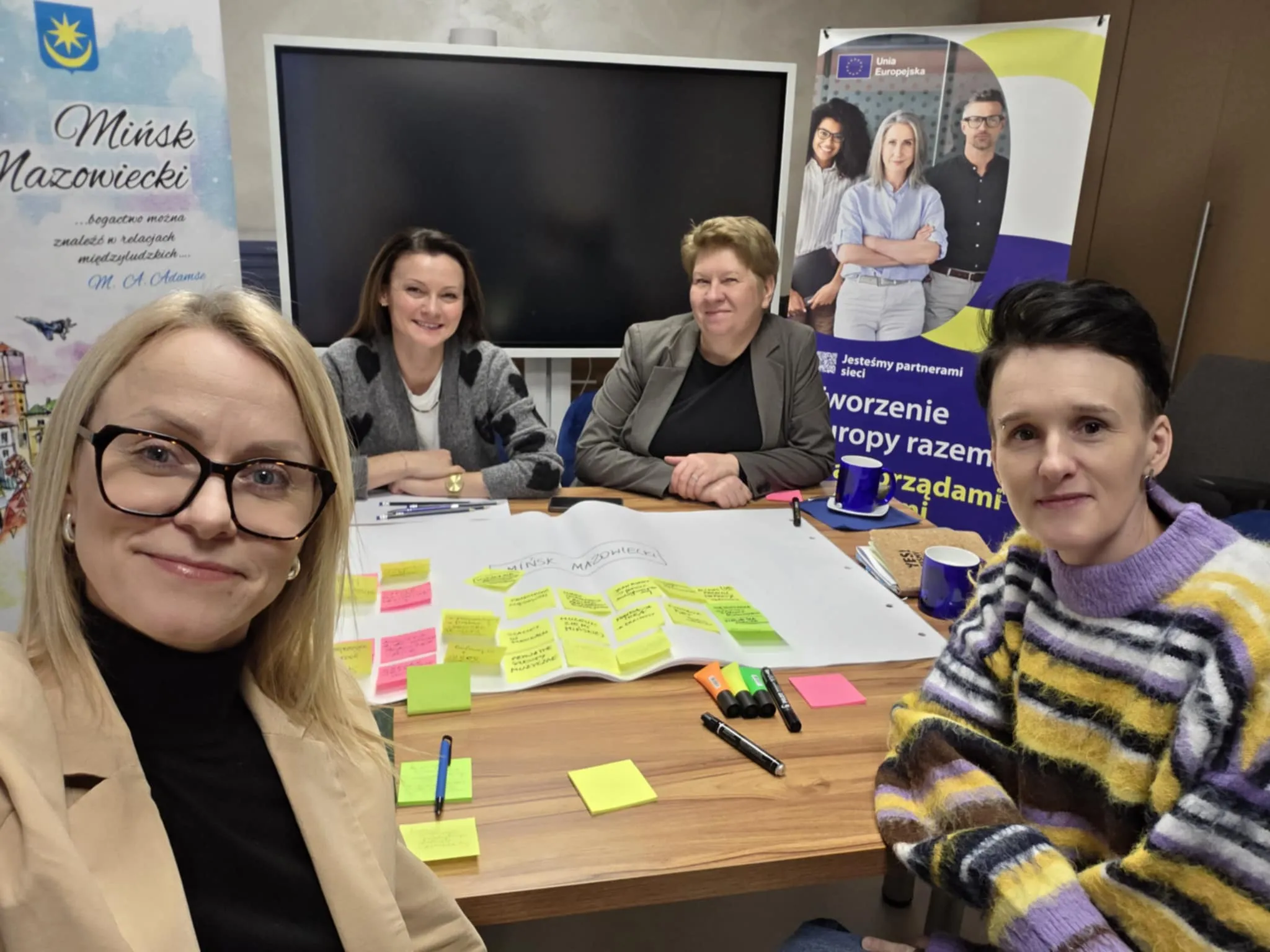 Cztery kobiety siedzą przy drewnianym stole w sali konferencyjnej. Kobieta z przodu po lewej robi selfie i ma okulary. Kobieta z przodu po prawej ma kolorowy pasiasty sweter. Dwie kobiety siedzą z tyłu przy stole. Na stole leży duży arkusz papieru z żółtymi, różowymi i zielonymi karteczkami samoprzylepnymi oraz flamastry i długopisy. W tle stoi duży czarny ekran i dwa stojące banery.