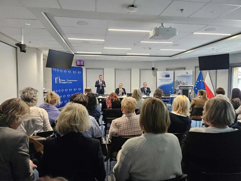 Grupa osób słuchająca trzech mężczyzn prowadzących spotkanie w sali konferencyjnej z flagami Unii Europejskiej i Polski.