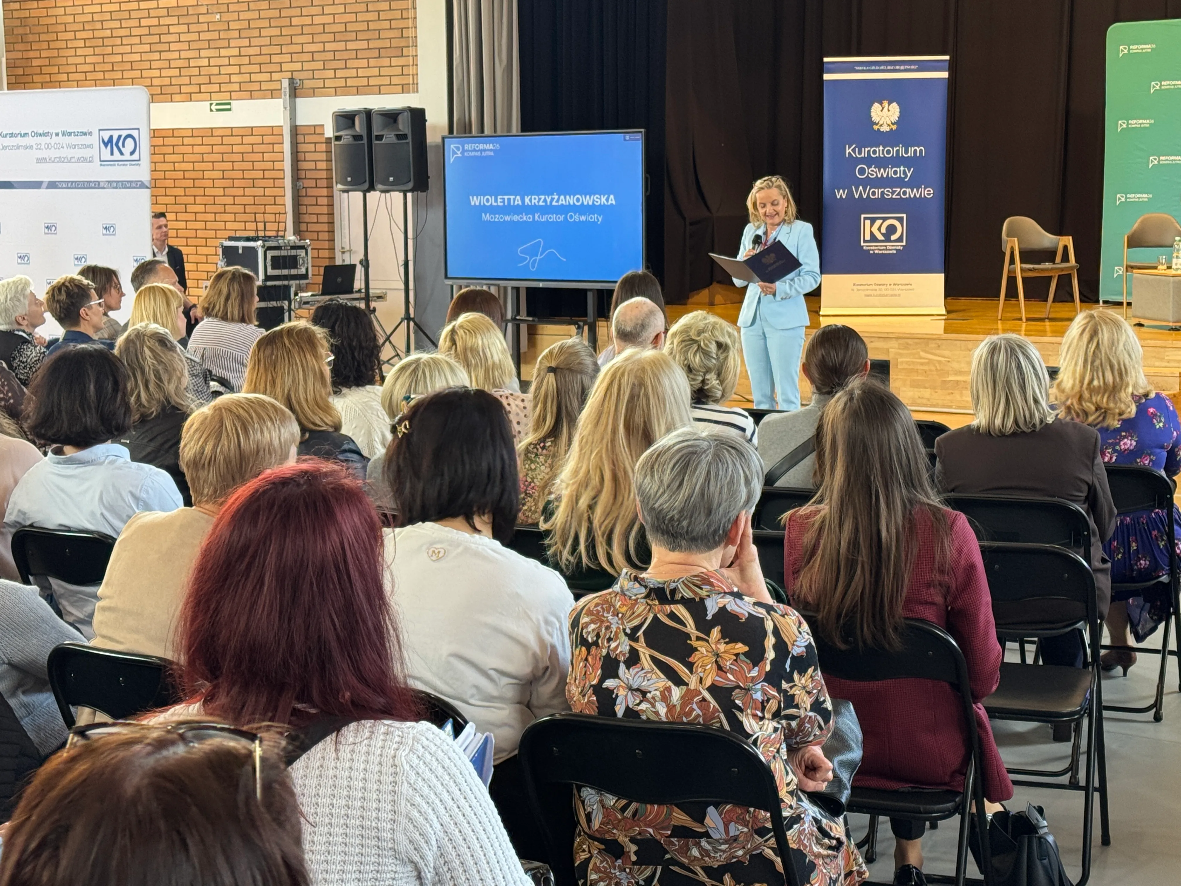 Publiczność siedzi na składanych krzesłach w sali z drewnianą sceną. Kobieta w jasnoniebieskim garniturze stoi na scenie i czyta z teczki. Obok stoi ekran z prezentacją i kilka banerów z herbem i logotypami. Widzowie to głównie dorośli, siedzą tyłem do aparatu.