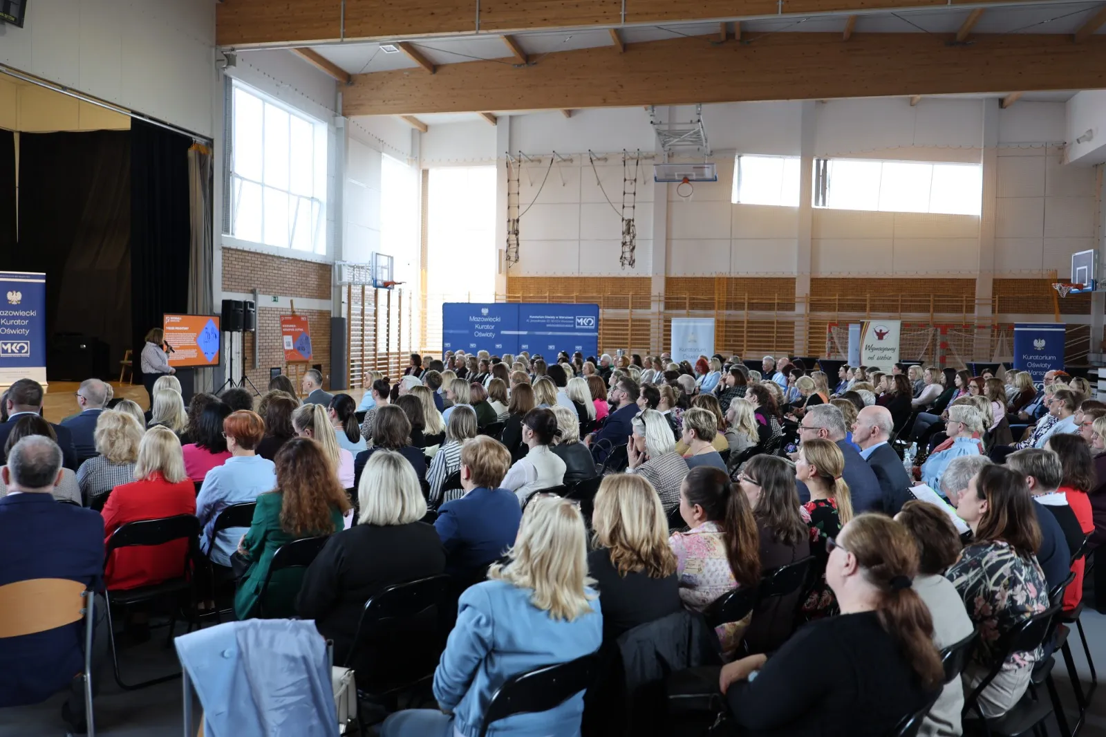 W sali gimnastycznej wiele osób siedzi w rzędach i słucha prelegentki przy mównicy. Jasne okna i drewniane belki sufitowe widoczne są nad aulą, z bocznymi banerami i koszem do koszykówki przy ścianie.
