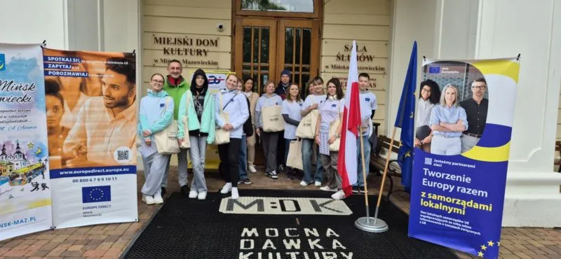 Grupa osób stojących na wejściu do budynku Miejskiego Domu Kultury w Mińsku Mazowieckim obok flag Polski i Unii Europejskiej oraz dwóch banerów informacyjnych.