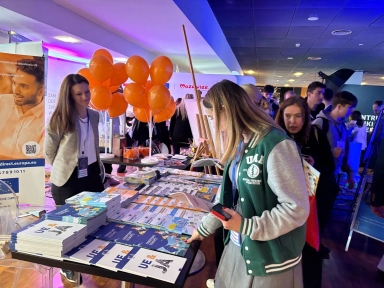 Wewnątrz budynku stoi kilka osób przy stoisku z ulotkami i książkami. Na stole są ułożone materiały promocyjne, obok znajduje się bukiet pomarańczowych balonów. W tle widoczne są kolejne osoby i inne stoiska, oświetlenie ma niebieską i fioletową barwę.