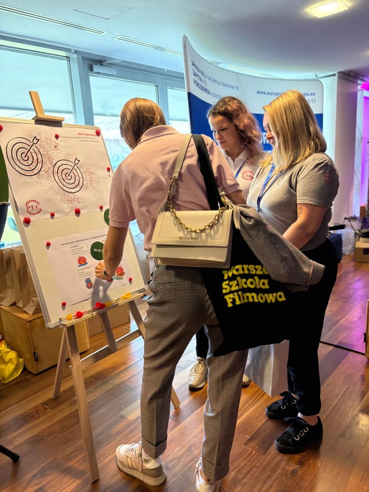 Trzy kobiety stoją przy planszy na sztaludze w jasnym pomieszczeniu z drewnianą podłogą. Jedna z nich wskazuje na planszę z wykresami i rysunkami tarcz ze strzałami. Kobiety mają na sobie casualowe ubrania, jedna z nich nosi torbę z napisem „warszawska szkoła filmowa”. W tle widoczny jest baner i okno.