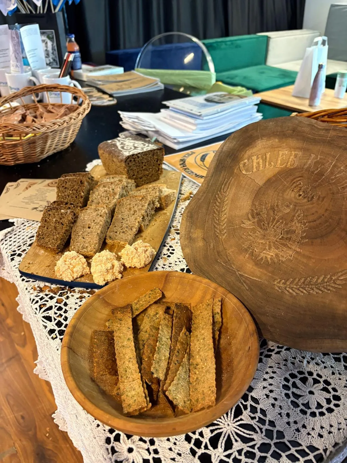 Na stole układane są kawałki ciemnego chleba na desce oraz chrupkie pieczywo w drewnianej misce. Obok leży drewniana okrągła deska z wyrytym wzorem i napisem. W tle widoczne są stosy książek, kubki, koszyk oraz meble z zielonym i jasnoszarym obiciem. Stół przykrywa biała koronkowa serweta.