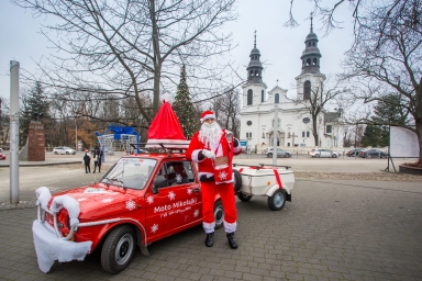 Mężczyzna w stroju Świętego Mikołaja stoi obok czerwonego samochodu ozdobionego na święta zimowe, z białym przyczepionym bokiem, na tle miejskiego placu z choinką i białym kościołem.