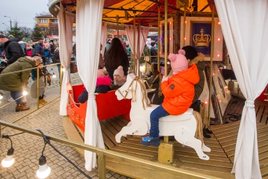 Dzieci bawiące się na karuzeli z drewnianymi konikami i czerwonym wózkiem. Karuzela znajduje się na zewnątrz, wśród zgromadzonych osób ubranych na zimę. W tle budynki i ozdobione światełkami drzewo. Wieczorna pora.