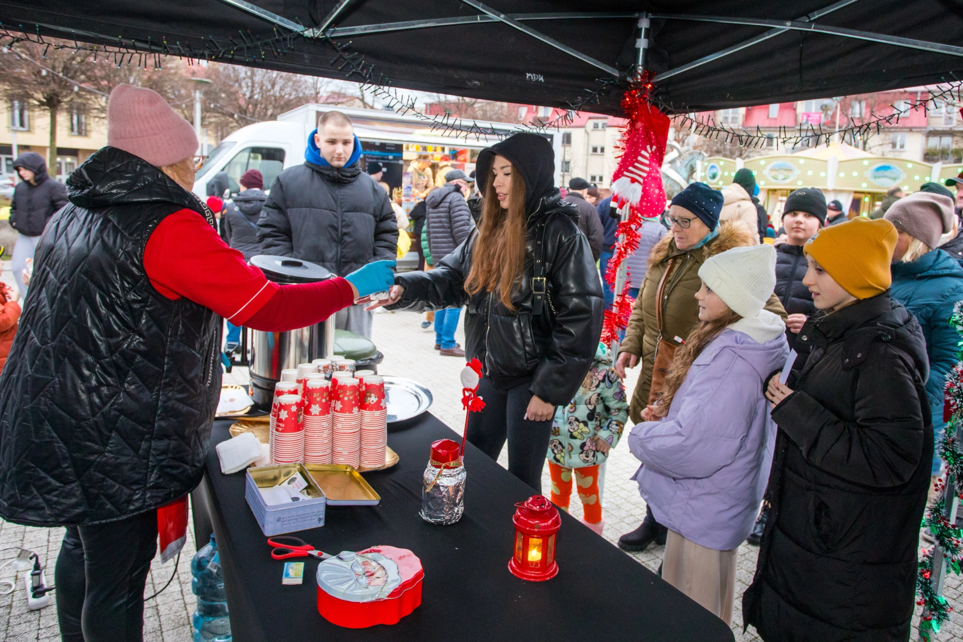 Stoisko na zewnątrz pod czarnym namiotem, kilka osób w zimowych ubraniach stoi w kolejce po napoje w papierowych kubkach z czerwonym świątecznym wzorem. Kobieta w różowej czapce i kamizelce podaje napój młodej kobiecie w czarnej kurtce z kapturem. Na stole są dekoracje świąteczne, w tym czerwony lampion i pudełko z Mikołajem. W tle widać więcej ludzi oraz budynki.