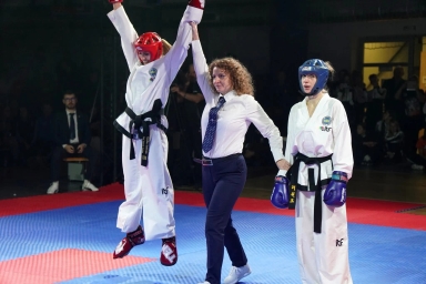 Sędzina podnosi rękę zawodniczki w czerwonym kasku, która skacze z radości, obok stoi zawodniczka w niebieskim kasku na niebiesko-czerwonej macie w hali sportowej.