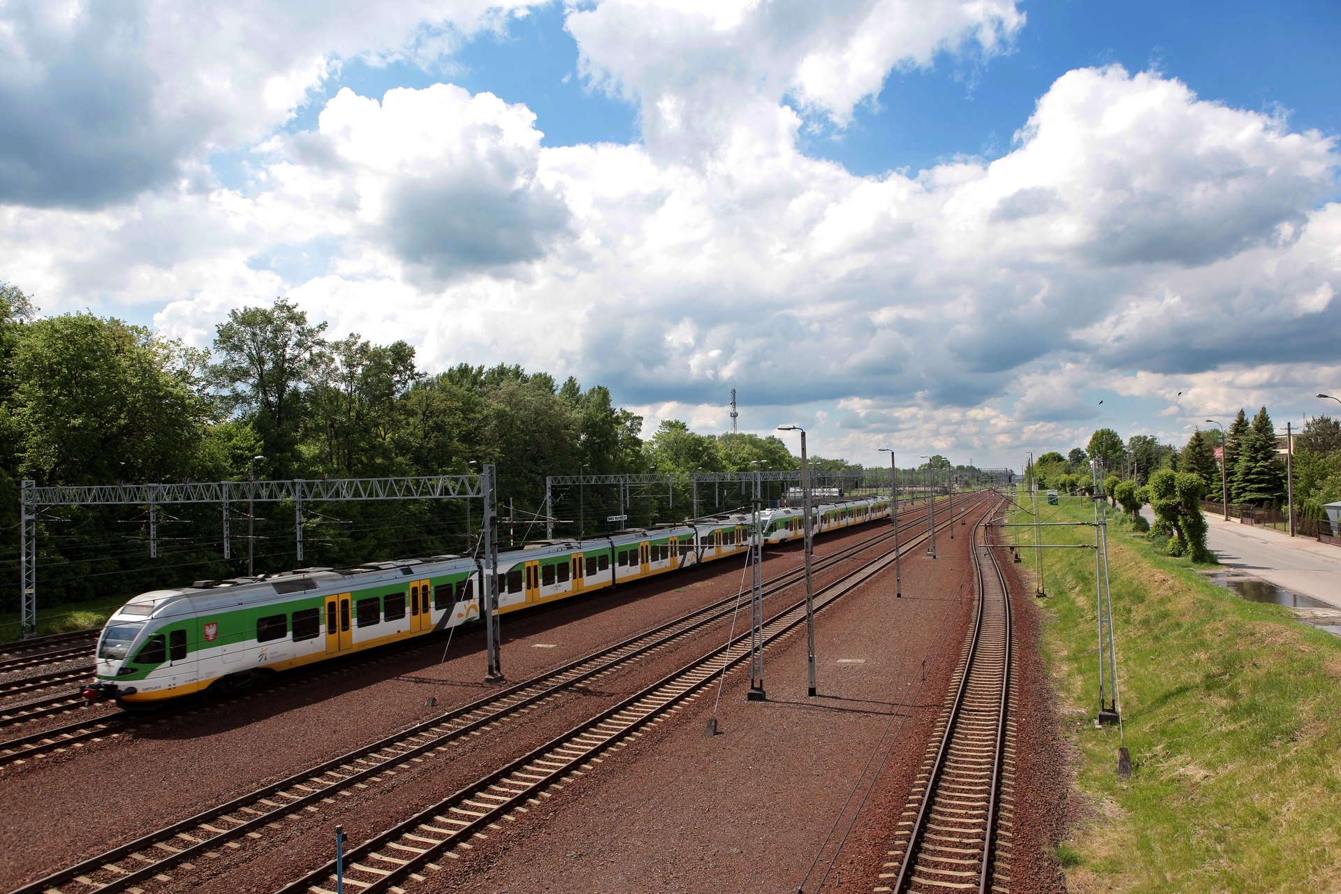 Pociąg osobowy w kolorach białym, zielonym i żółtym na torach kolejowych obok zielonych drzew i drogi.