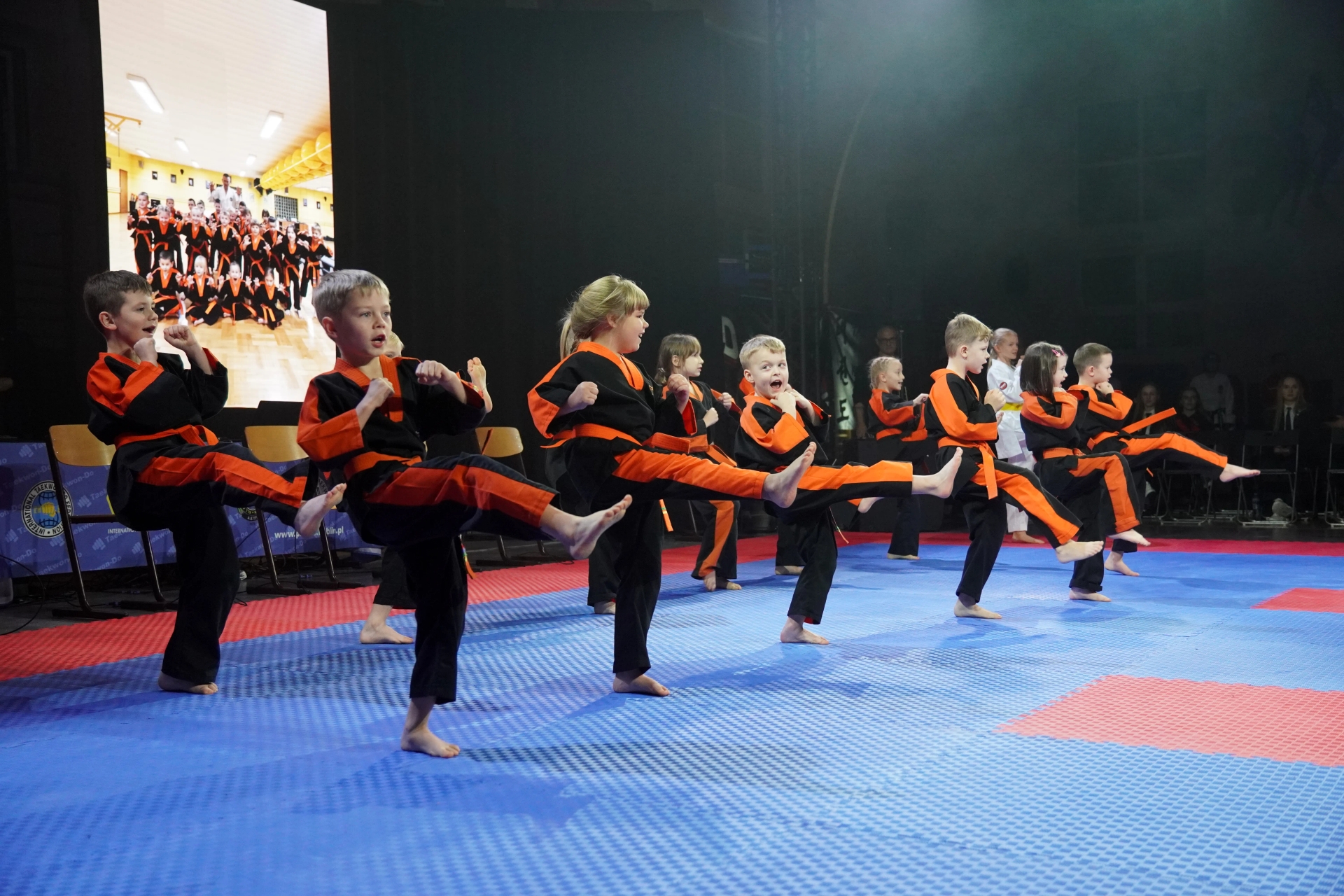 Dziesięcioro dzieci ćwiczy kopnięcia w stroju do karate o czarno-pomarańczowym kolorze na niebiesko-czerwonej macie, w tle ciemne wnętrze sali i ekran z grupowym zdjęciem dzieci w tych samych strojach.