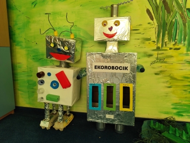 Dwa roboty z kartonów i folii aluminiowej stoją wewnątrz przed jasną zieloną ścianą. Mniejszy po lewej ma srebrną głowę z czerwonym uśmiechem, biały tułów ozdobiony plastikowymi nakrętkami i kartonowymi elementami oraz nogi owinięte folią stojące na opakowaniach na jajka. Większy po prawej ma prostokątny srebrny korpus z trzema pionowymi otworami do segregacji, cylindryczne stopy i butelki jako ramiona.