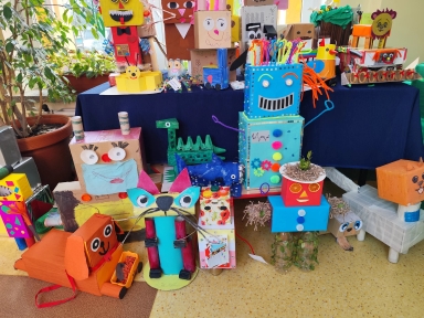 Kolorowe postacie z kartonów i plastikowych butelek stoją i leżą w sali przy stole z granatowym obrusem. W centrum stoi wysoki niebieski robot z pomarańczowymi drucikowymi włosami i dużym uśmiechem. Po lewej roślina doniczkowa przy oknie i kilka papierowych figurek. Na podłodze leży pomarańczowy pies z pudełka i kot z dużymi oczami. Jasna podłoga, wnętrze, dzień.