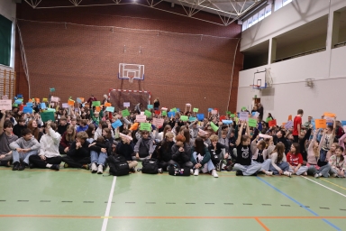 Grupa dzieci i młodzieży siedzi na podłodze hali sportowej. Trzymają kolorowe kartki uniesione do góry. W tle ceglana ściana z bramką do piłki ręcznej i kosz do koszykówki.