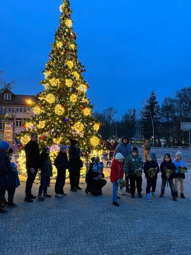 Grupa dzieci i dorosłych stoi na zewnątrz przy wysokiej, ozdobionej świątecznej choince z jasnymi żółtymi i czerwonymi dekoracjami, widoczne wieczorne, niebieskie niebo. Dzieci trzymają pluszowe zabawki. W tle budynki i drzewa bez liści.