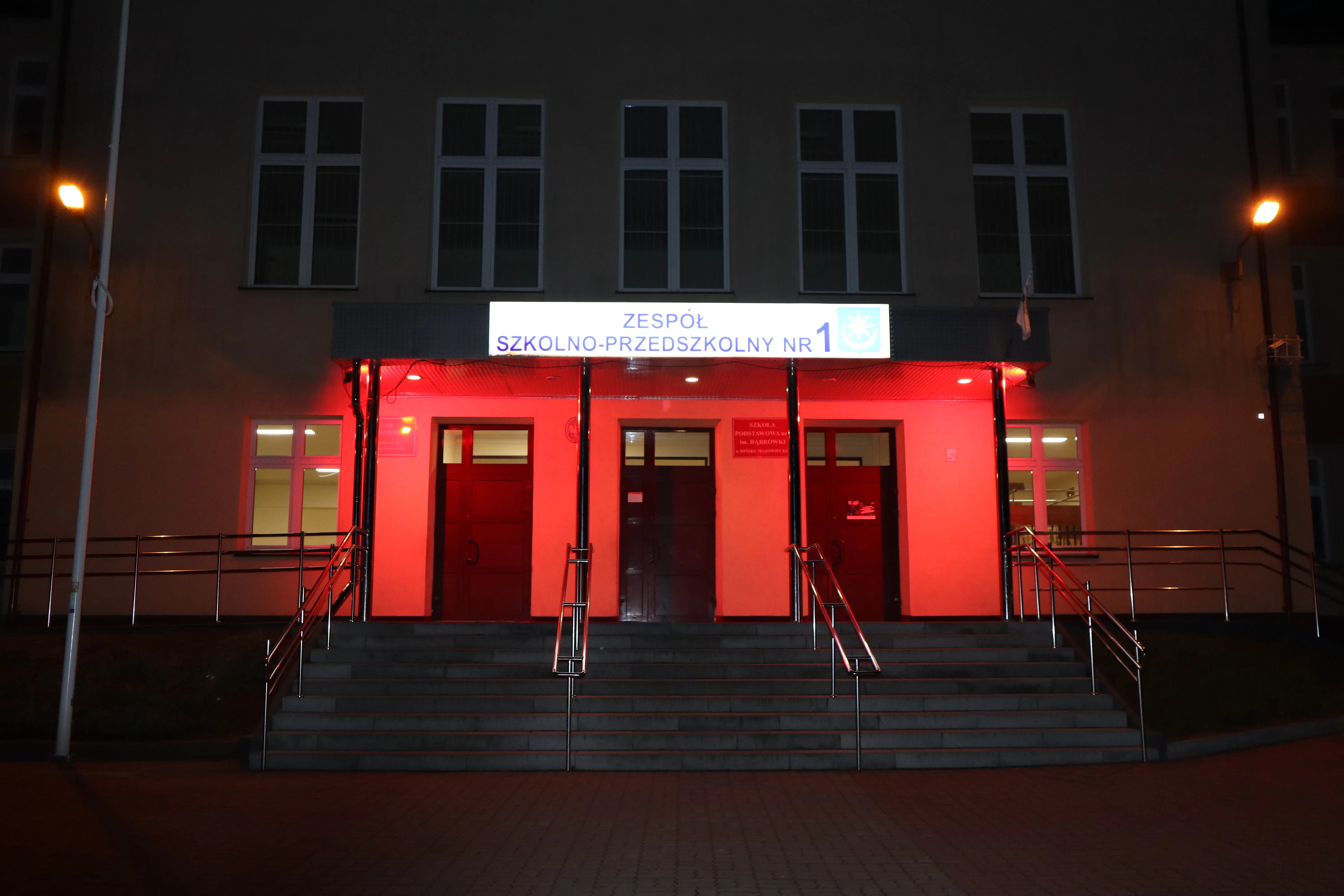 Wejście do budynku szkoły z trzema drzwiami i schodami prowadzącymi do nich, oświetlone na czerwono, z jasną tablicą informacyjną nad drzwiami, widoczne dwa lampy uliczne po bokach.
