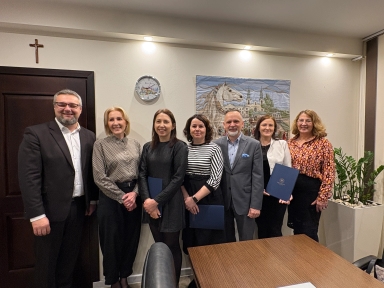 Siedem osób stoi obok siebie w pomieszczeniu biurowym przy jasnej ścianie z obrazem i zegarem. Trzy osoby trzymają w rękach teczki. Po prawej stronie widać dużą donicę z rośliną. Na górze po lewej stronie na ścianie wisi krzyż.