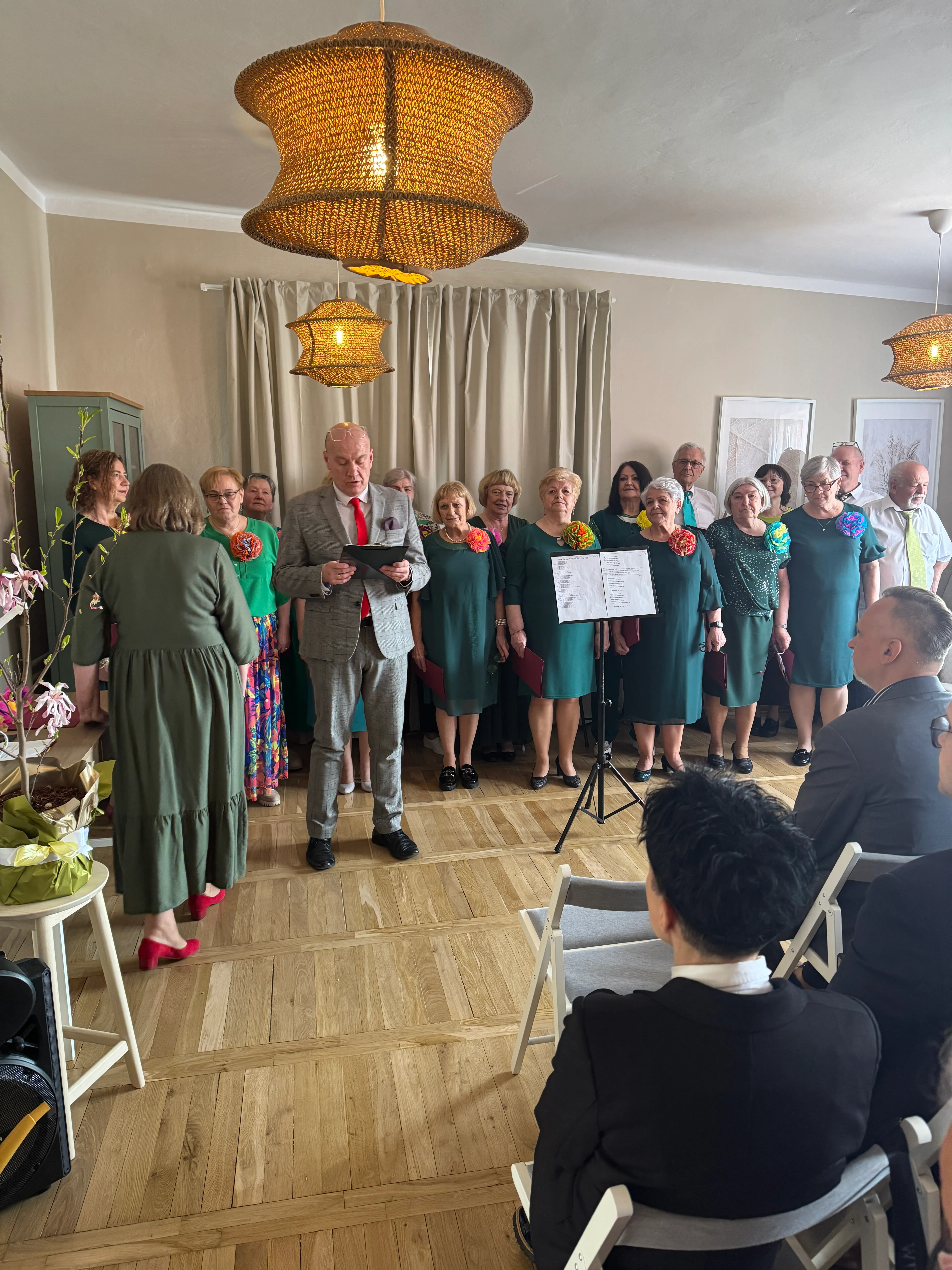 Grupa kilkunastu dorosłych stoi w rzędzie wewnątrz pokoju i śpiewa. Mężczyzna w szarej marynarce z czerwonym krawatem stoi z przodu i czyta z kartki. Kobiety w zielonych sukienkach noszą duże kolorowe kwiaty przypięte przy ramieniu. Na środku stoi pulpit z nutami. Pomieszczenie ma drewnianą podłogę, beżową zasłonę na tle i trzy wiszące plecione lampy dające ciepłe światło. Na pierwszym planie siedzą widzowie na składanych krzesłach.