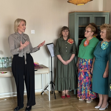 Kobieta po lewej stoi i gestykuluje mówiąc do grupy. Po prawej trzy starsze kobiety stoją obok siebie w kolorowych sukienkach z dużymi broszami. Wnętrze z jasną ścianą, zieloną szafką, stołem z butelkami wody i pulpitowym stojakiem na nuty.