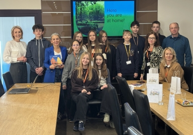 Grupa piętnastu osób, w tym młodzież i dorośli, stoi i siedzi przy stole konferencyjnym w jasnym pomieszczeniu biurowym. Na ścianie za nimi znajduje się ekran z napisem „Here you are at home!” na zielonym tle z widokiem na park. Na stole leżą torby papierowe, laptop i przekąski.
