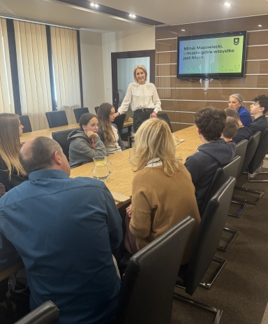 Grupa dziesięciu osób siedzi wokół drewnianego stołu w sali konferencyjnej, jedna kobieta stoi i przemawia. Na ścianie widoczny jest ekran z napisem „Mińsk Mazowiecki, miasto gdzie wszystko jest blisko”. Wnętrze jest jasno oświetlone, okna zasłonięte roletami.