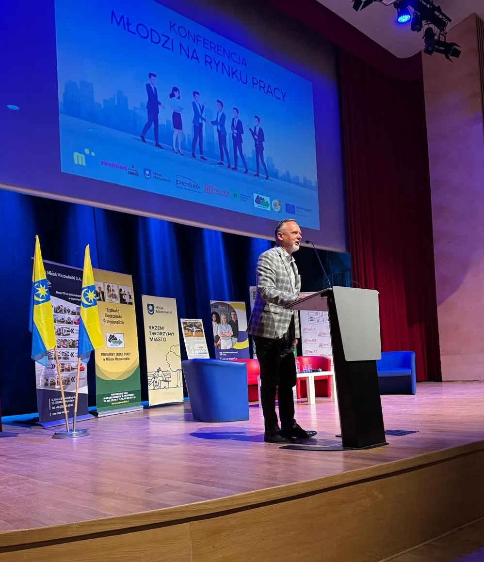 Mężczyzna stoi przy pulpicie na scenie i przemawia. Sala jest wewnątrz, scena ma niebieskie oświetlenie. Za mówcą wisi duży ekran z ilustracją sylwetek biznesowych. Po lewej stoją banery i dwie żółto-niebieskie flagi, na scenie są też niebieskie i czerwone fotele.