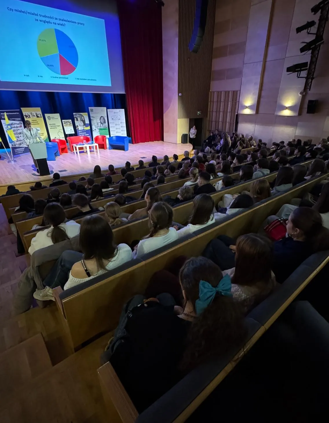 Wnętrze audytorium. Publiczność liczy kilkadziesiąt osób i siedzi na tarasowo ułożonych drewnianych ławkach. Na scenie po lewej stoi mówca przy mównicy. Na scenie są czerwone i niebieskie fotele oraz niski stolik. Za sceną wisi duży ekran z wyświetloną kolorową grafiką i czerwone zasłony. Sala jest oświetlona sztucznym światłem i ma drewniane panele ścienne.