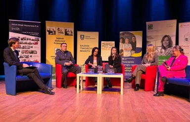 Sześć osób siedzi na scenie w kolorowych fotelach podczas panelu dyskusyjnego. Przed nimi stoi niski stolik z dzbankiem wody i szklankami. Za nimi wiszą pionowe banery reklamowe na żółtym i niebieskim tle. Scena ma drewnianą podłogę i niebieskie oświetlenie.