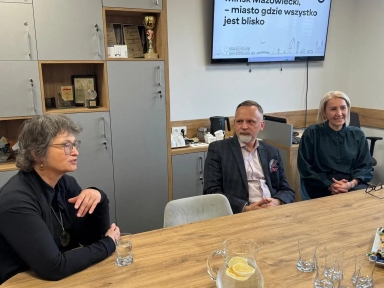 Jasne pomieszczenie. Trzy osoby siedzą przy drewnianym stole w sali konferencyjnej. Po lewej kobieta z krótkimi szarymi włosami i okularami, pośrodku mężczyzna w marynarce w kratę, po prawej kobieta z blond włosami ubrana na czarno. Na stole dzbanek z wodą i plasterkami cytryny oraz puste szklanki. W tle znajdują się szare szafki, na ścianie zawieszony ekran