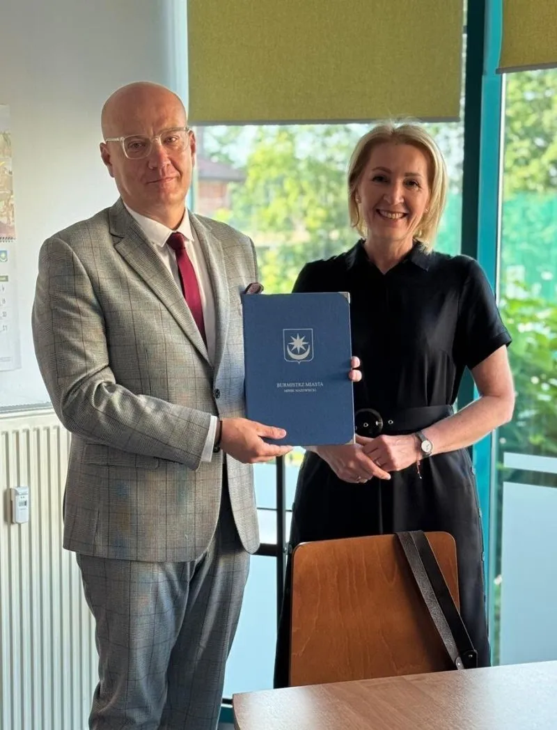 Mężczyzna w szarym garniturze trzyma granatową teczkę. Stoi obok uśmiechającej się kobiety w czarnej sukience.