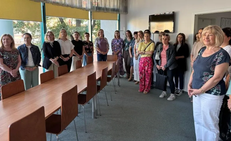 Grupa kobiet stoi wzdłuż ściany i długiego stołu w jasnym pomieszczeniu z dużymi oknami.