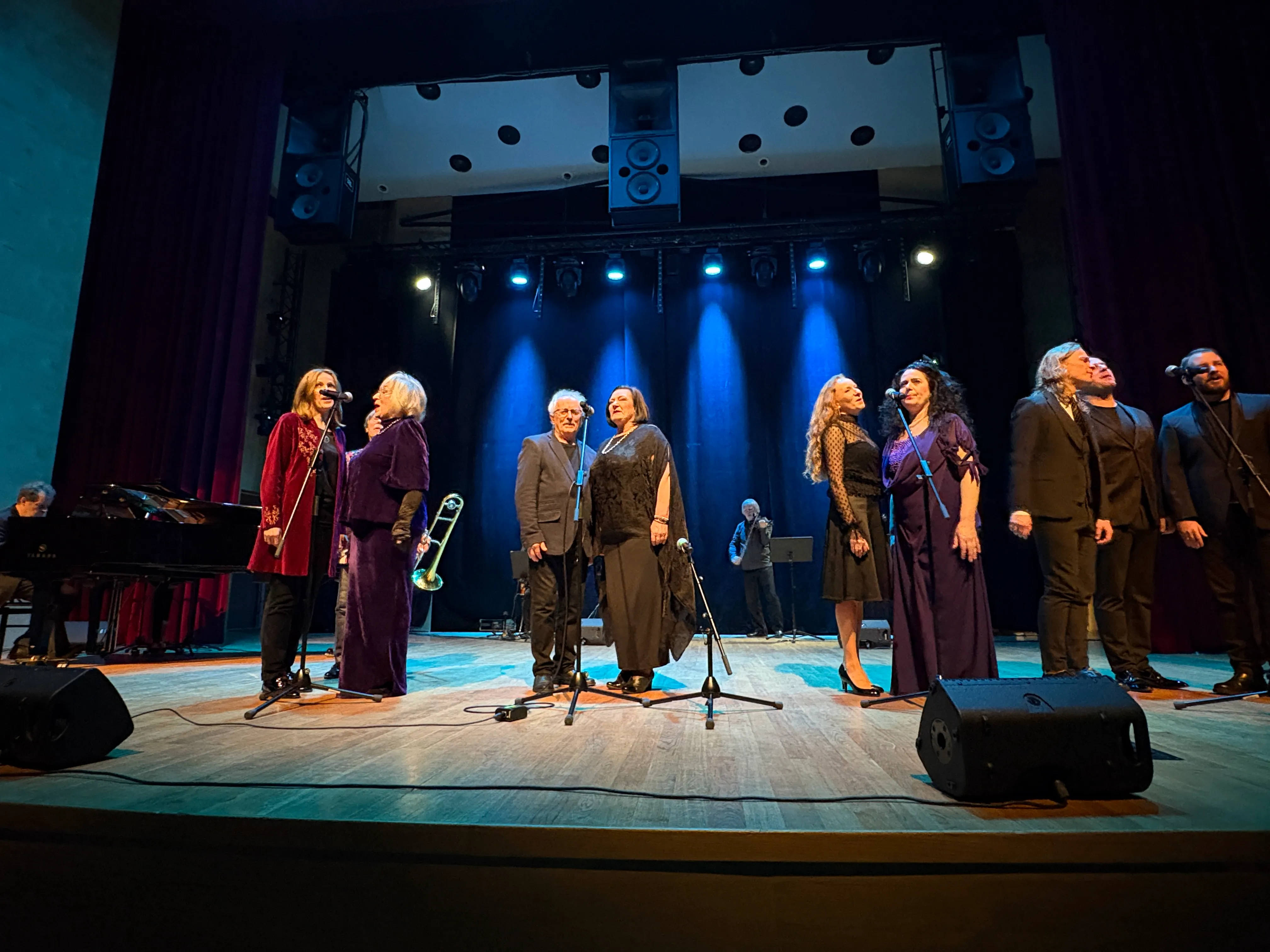 Dziewięć osób stoi na oświetlonej scenie teatru, w tle ciemna kurtyna i niebieskie światła. Po lewej stronie mężczyzna gra na fortepianie, druga osoba ma puzon. Kilka osób stoi przy mikrofonach, ubranych w eleganckie stroje.