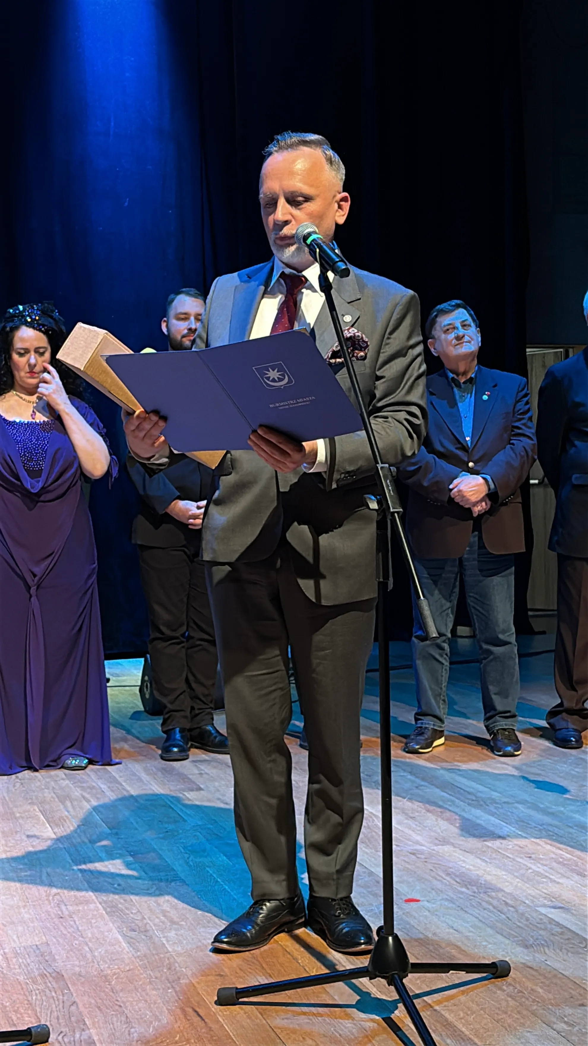 Mężczyzna w szarym garniturze stoi przy mikrofonie na scenie, czytając z niebieskiego folderu. Za nim stoi pięć osób w formalnych strojach, dwie kobiety i trzej mężczyźni. Scena jest oświetlona niebieskim światłem, a tło jest ciemne.