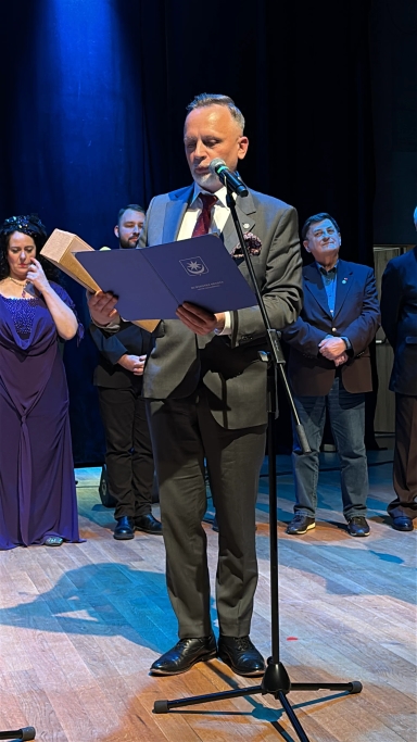 Mężczyzna w szarym garniturze stoi przy mikrofonie na scenie, czytając z niebieskiego folderu. Za nim stoi pięć osób w formalnych strojach, dwie kobiety i trzej mężczyźni. Scena jest oświetlona niebieskim światłem, a tło jest ciemne.