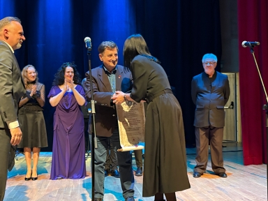 Sześć osób na scenie podczas ceremonii wręczenia dyplomu. Mężczyzna w ciemnej marynarce podaje dyplom kobiecie w czarnej sukience. Trzy osoby z tyłu klaszczą, jedna stoi obok mikrofonu po prawej stronie. Tło sceny jest ciemnoniebieskie, z prawej strony czerwona kurtyna.