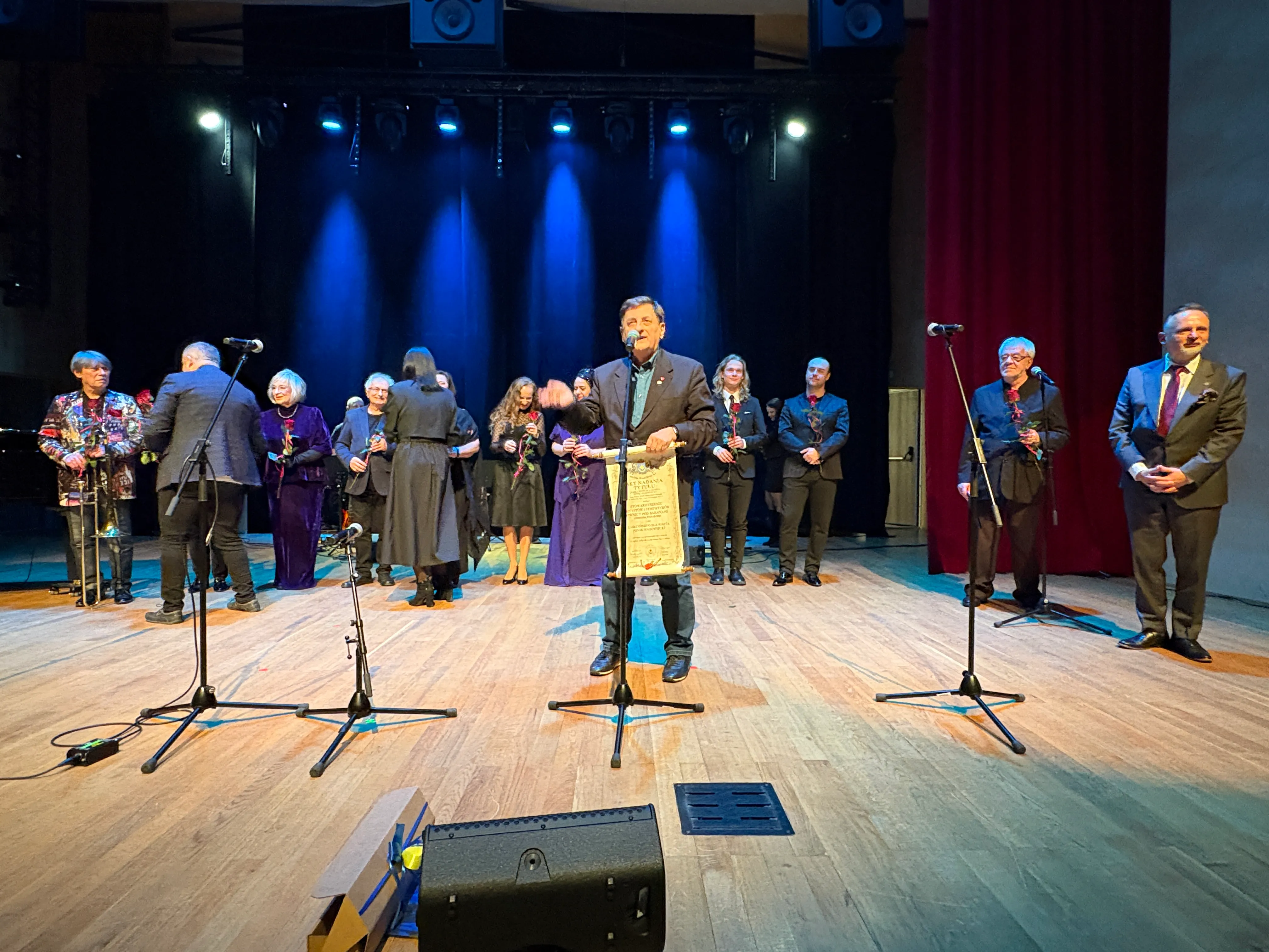 Grupa około piętnastu osób stoi na scenie z drewnianą podłogą pod niebieskim oświetleniem. Mężczyzna na środku przemawia do mikrofonu, trzymając zwój. Inni trzymają czerwone róże, ubrani są w eleganckie stroje. Z boku sceny wiszą czerwone kotary.