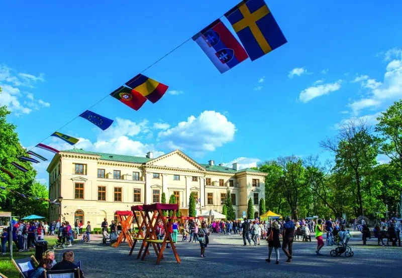 Plac z tłumem ludzi i drewnianymi konstrukcjami przed klasycystycznym budynkiem z flagami państw powiewającymi na linie nad placem.