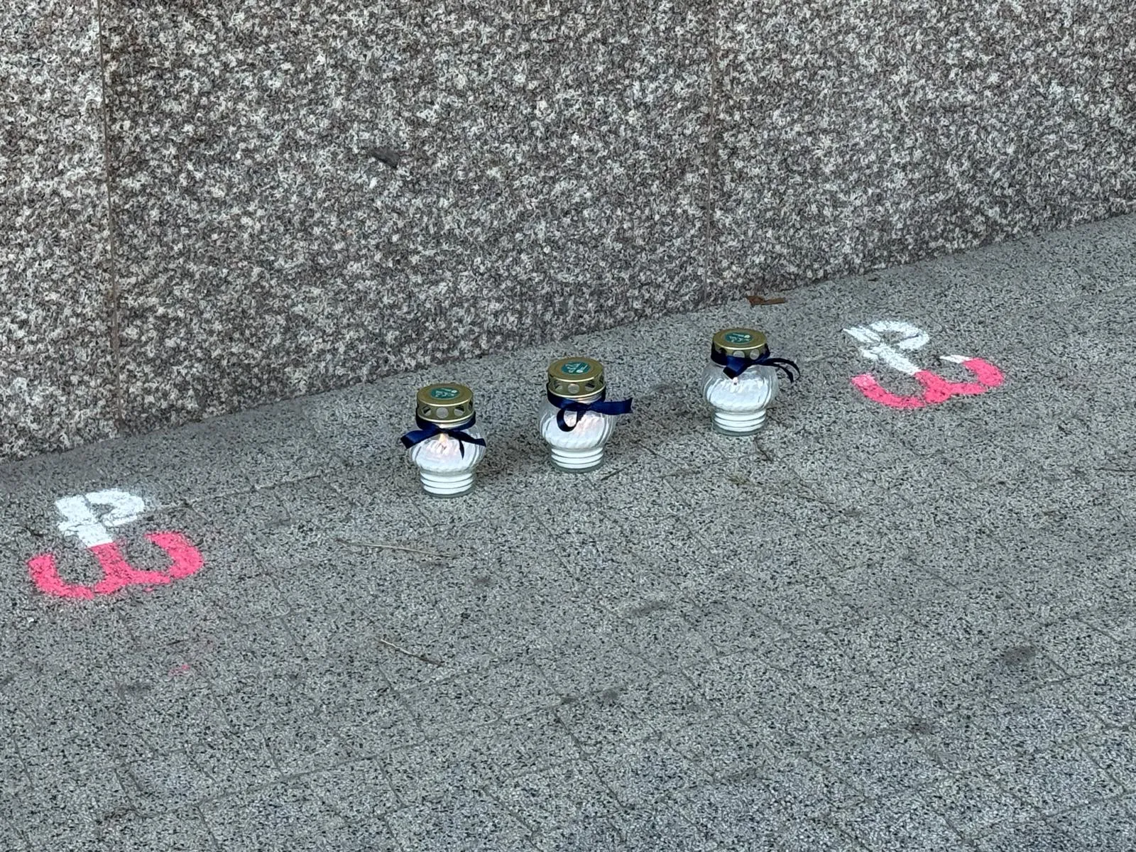 Trzy znicze ustawione na szarym chodniku przy ścianie z granitu, obok nich na chodniku namalowane symbole Polski Walczącej.